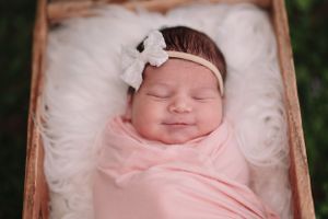 Sleeping newborn swaddled in soft pink wrap with white bow headband resting in wooden crate.