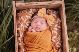 Peacefully sleeping newborn wrapped in mustard yellow swaddle with matching bow in wooden box.
