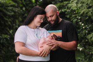 Parents share tender outdoor moment while holding their newborn baby among green foliage.