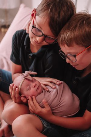 Two siblings gently holding and admiring a sleeping newborn baby wearing pink.
