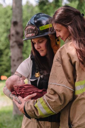 Firefighters in protective gear tenderly cradling newborn wrapped in burgundy cloth outdoors.