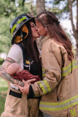 Firefighter couple in uniform sharing tender moment while holding their newborn outdoors.