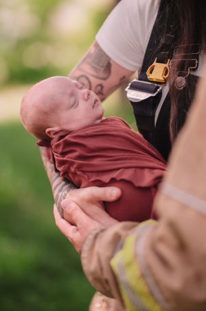 Close-up of newborn in burgundy wrap being held by someone in firefighter gear.