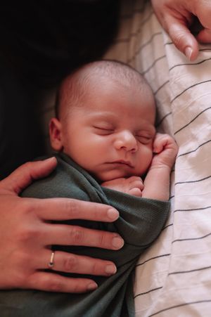 Sleeping newborn in grey outfit nestled against striped fabric with gentle hands holding.