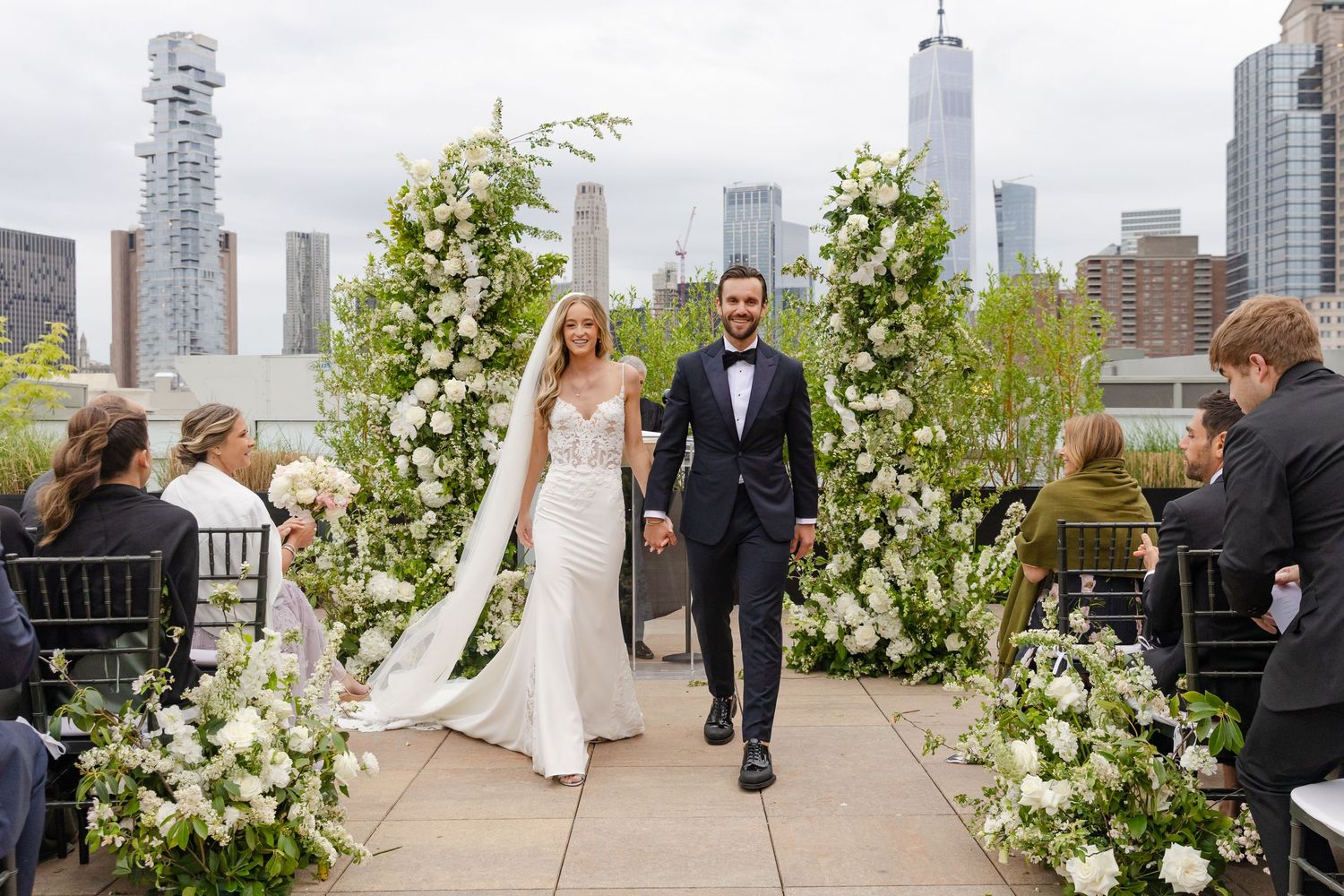 Elegant white floral arch and greenery decorate an outdoor rooftop wedding ceremony space with city skyline views.
