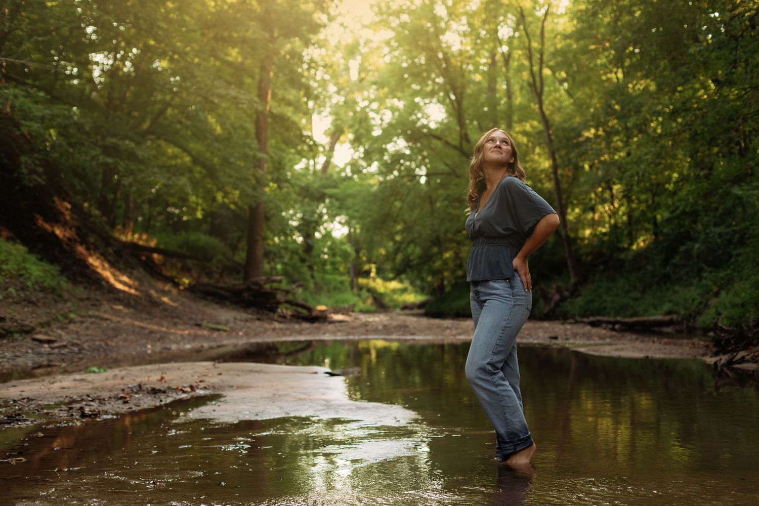 Neoga senior girl stands in shallow creek with blue jeans and pretty blue blouse.