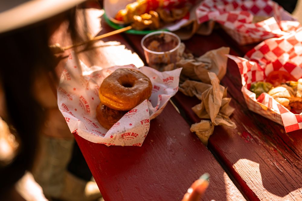 A glazed donut and fried foods served in red checkered paper baskets on a rustic wooden picnic table outdoors.