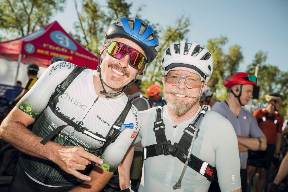 Two smiling cyclists in helmets and gear pose together at an outdoor cycling event on a sunny day.