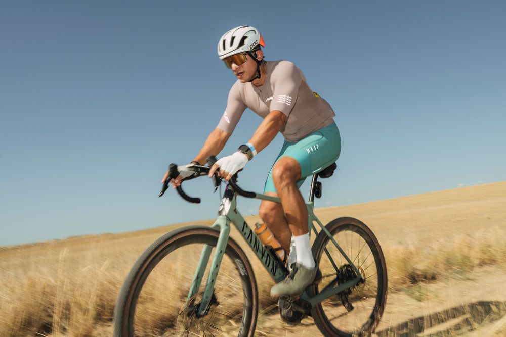 Cyclist riding a green gravel bike through open fields wearing a white helmet and teal shorts on a sunny day.