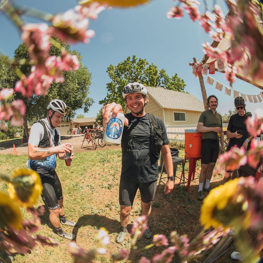 Cyclists in helmets smile at a sunny outdoor rest stop with pink flowers, bunting flags, and a red grill nearby.