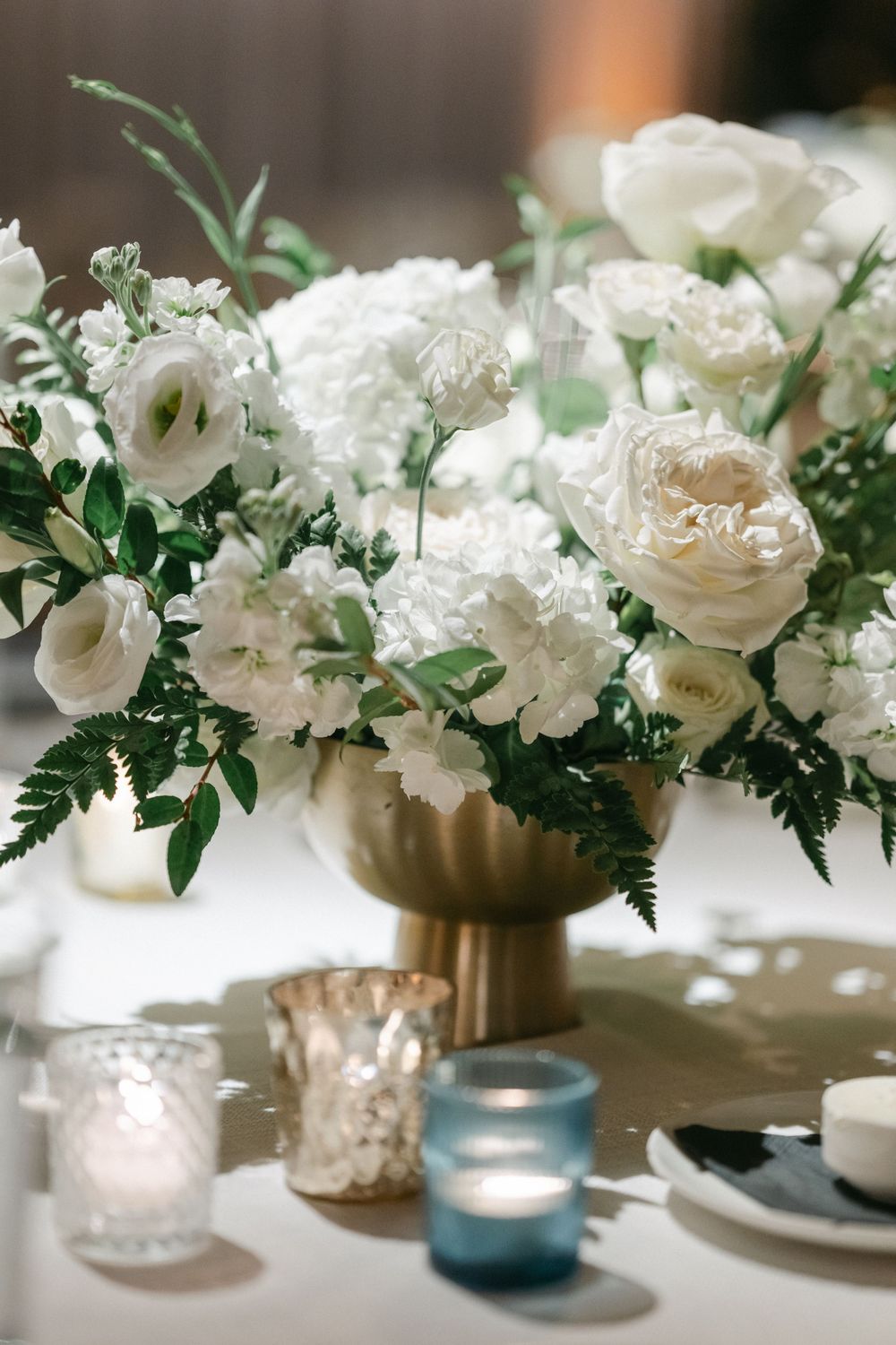Elegant white floral centerpiece with roses and greenery in gold vase alongside blue and mercury glass votives on white tablecloth. - RPM