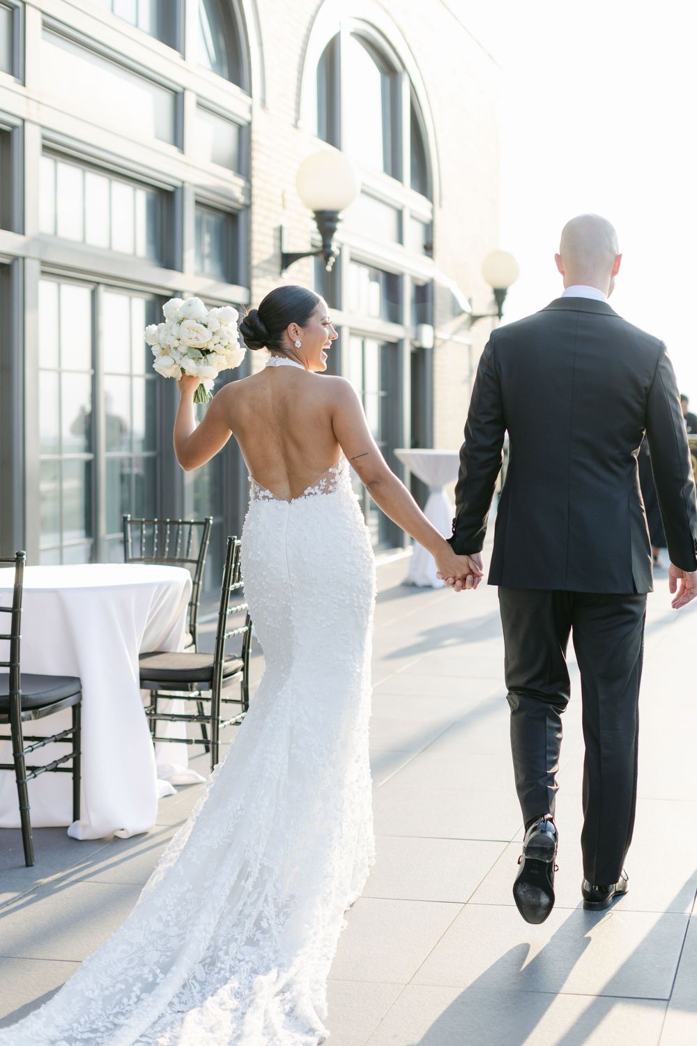 Bride and groom walk hand in hand along a sunlit building exterior with white rocking chairs and windows. - Penthouse Hyde Park