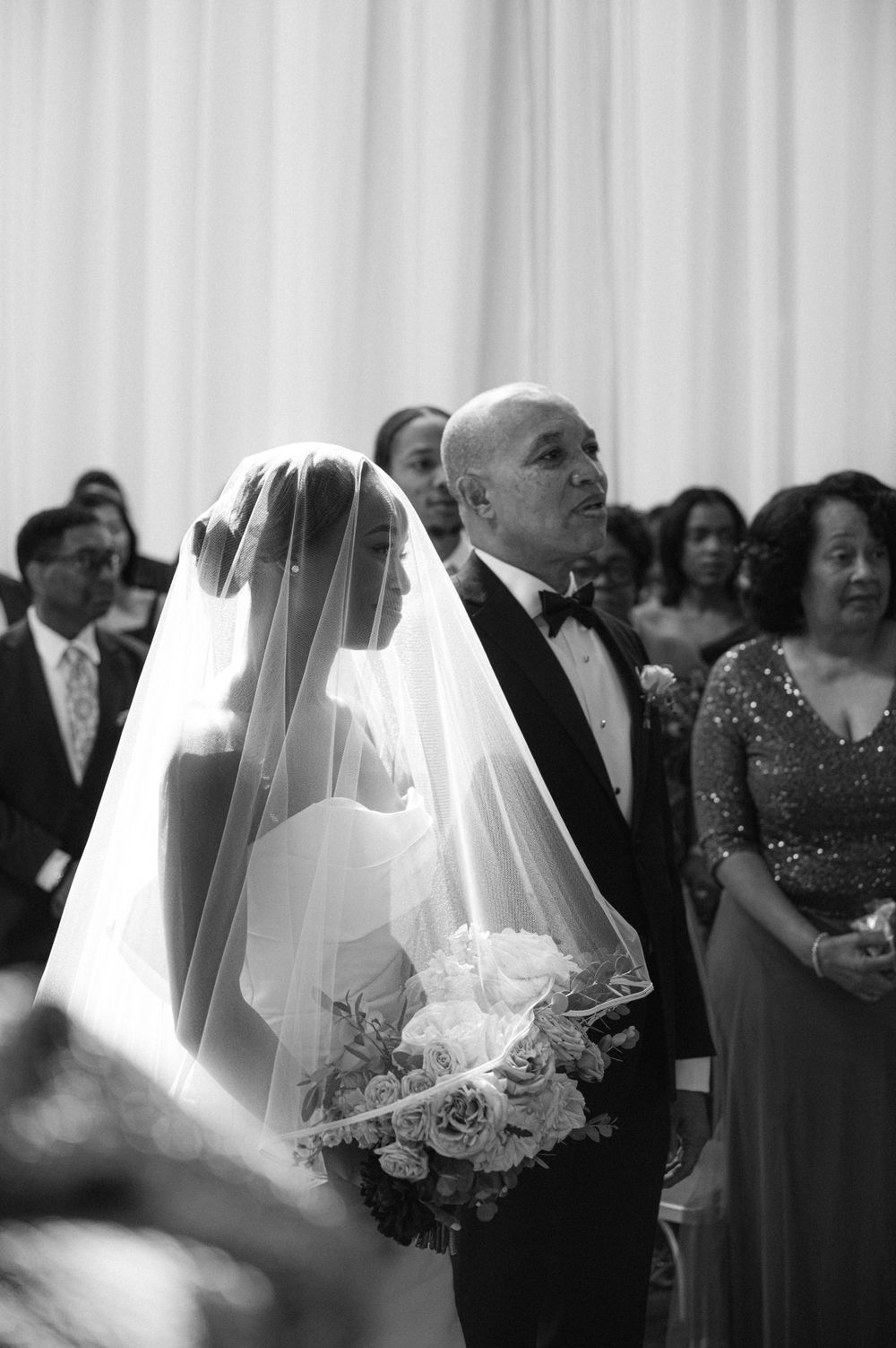 Black and white candid wedding ceremony photo showing bride with veil and guests in background.