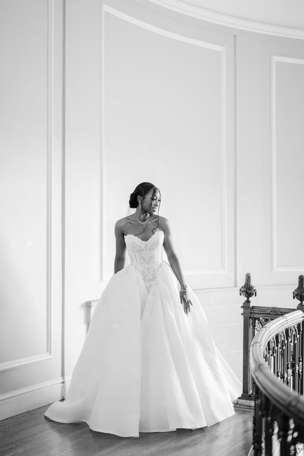 A stunning black and white photograph of a bride in an elegant strapless ball gown standing by a staircase. - The Armour House Wedding