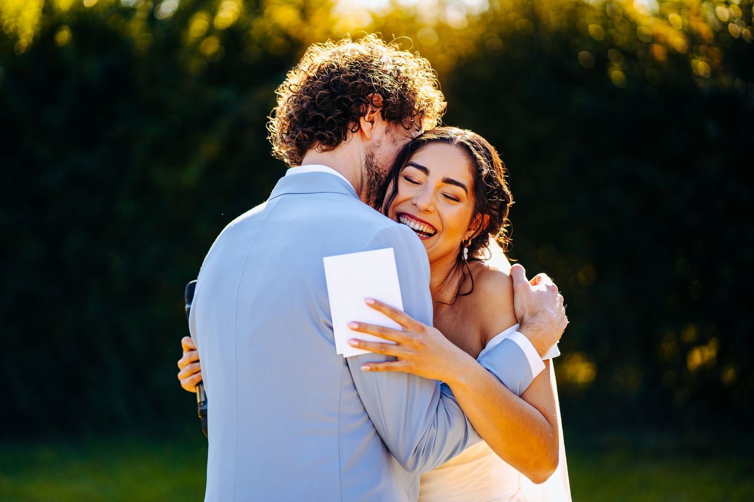 Bride and groom share first kiss at outdoor wedding ceremony surrounded by cheering guests in colourful attire.