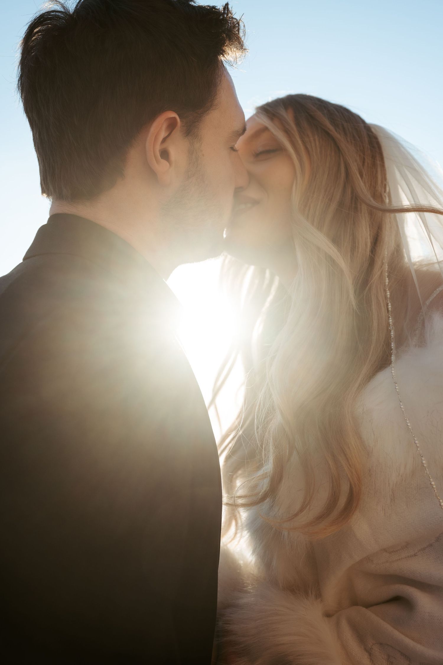 Happy couple in wedding attire playfully dance on a porch near a 'Your Spot' sign in sunlit outdoor setting.