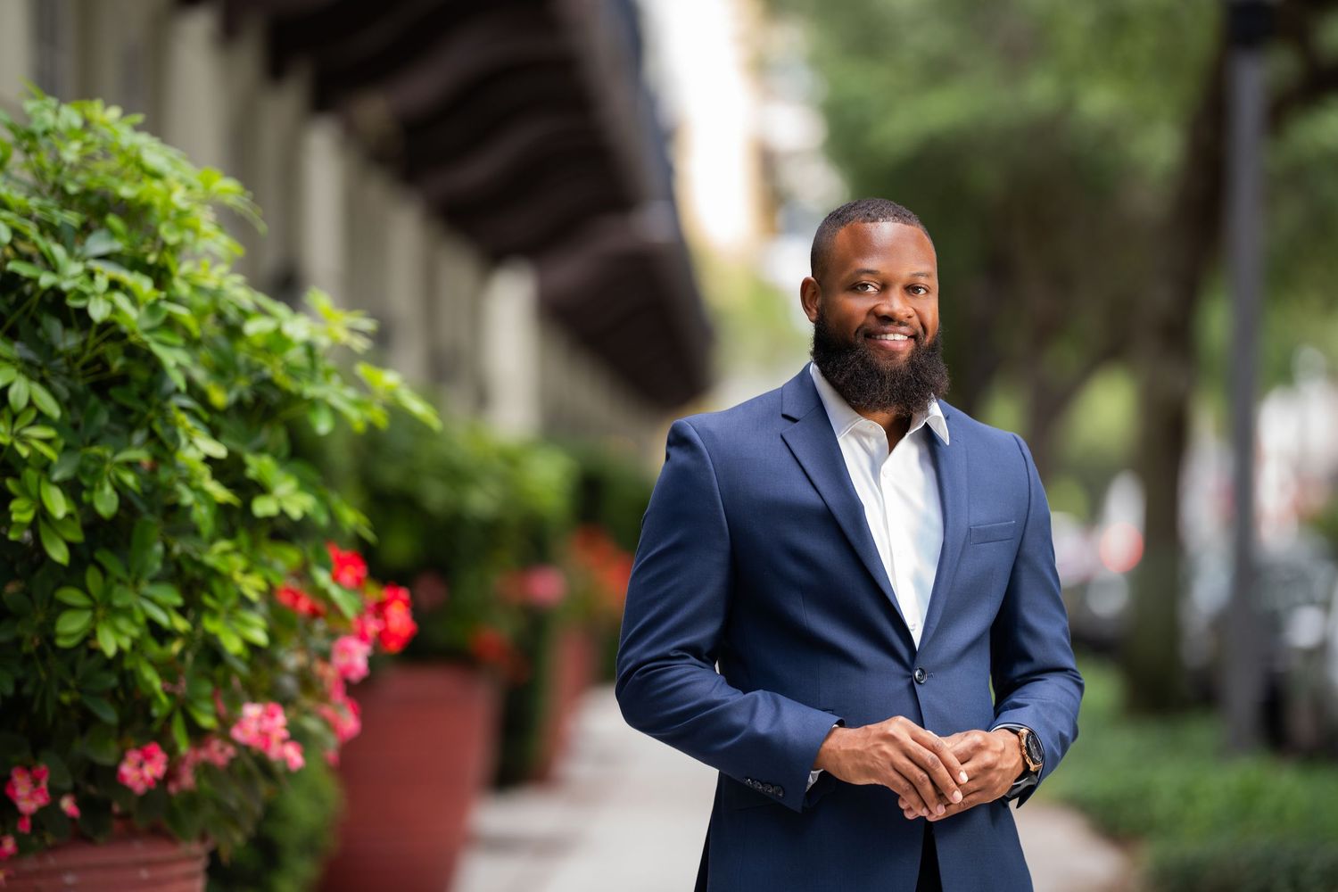 Professional in a navy blue suit stands smiling outdoors near greenery and pink flowers on an urban street.