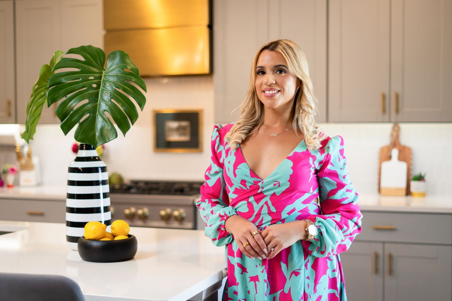 A person in a vibrant pink and blue floral dress stands in a modern kitchen with a monstera plant in striped vase.