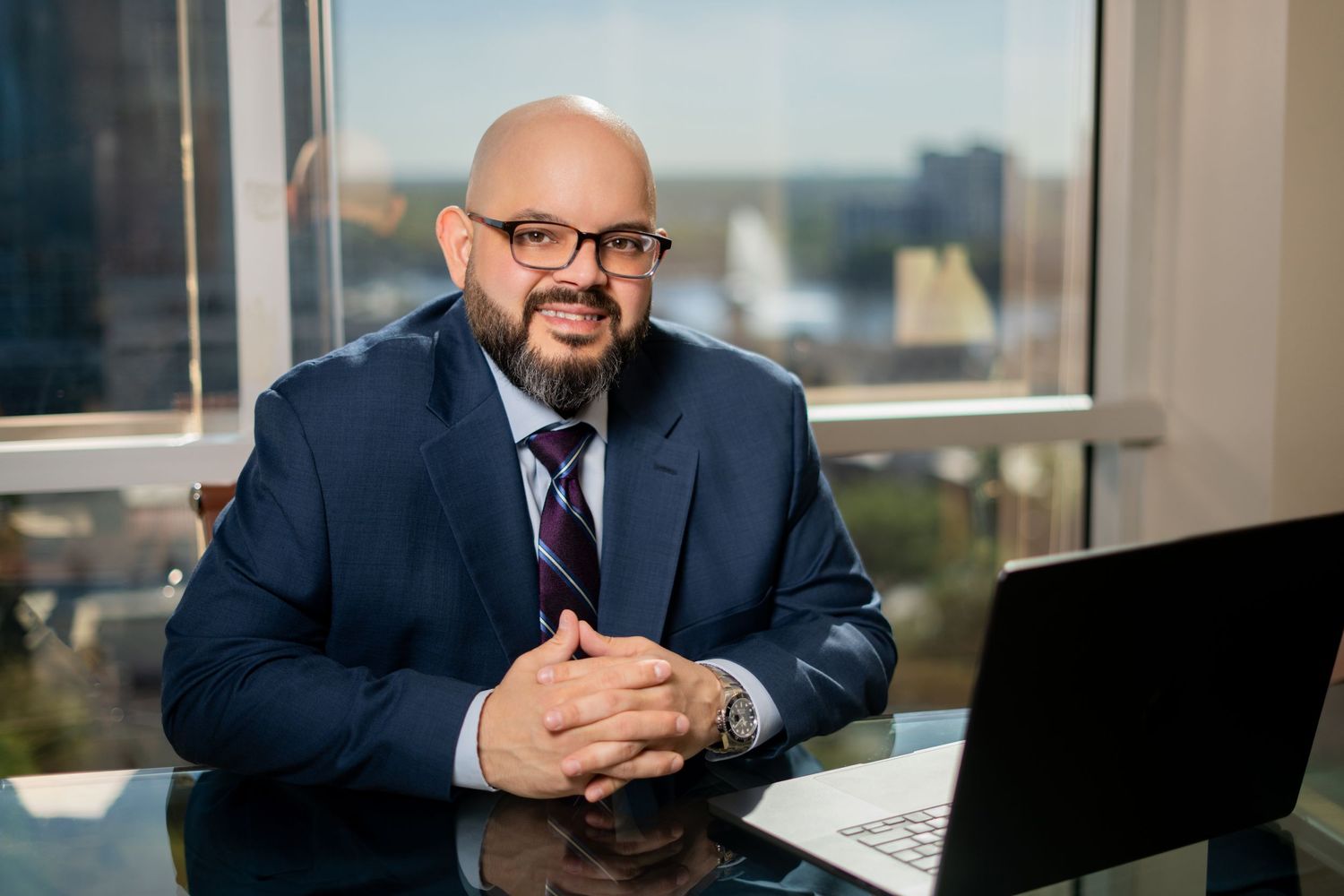 Professional in a navy suit and striped tie sits at desk with laptop in modern office with city view background.