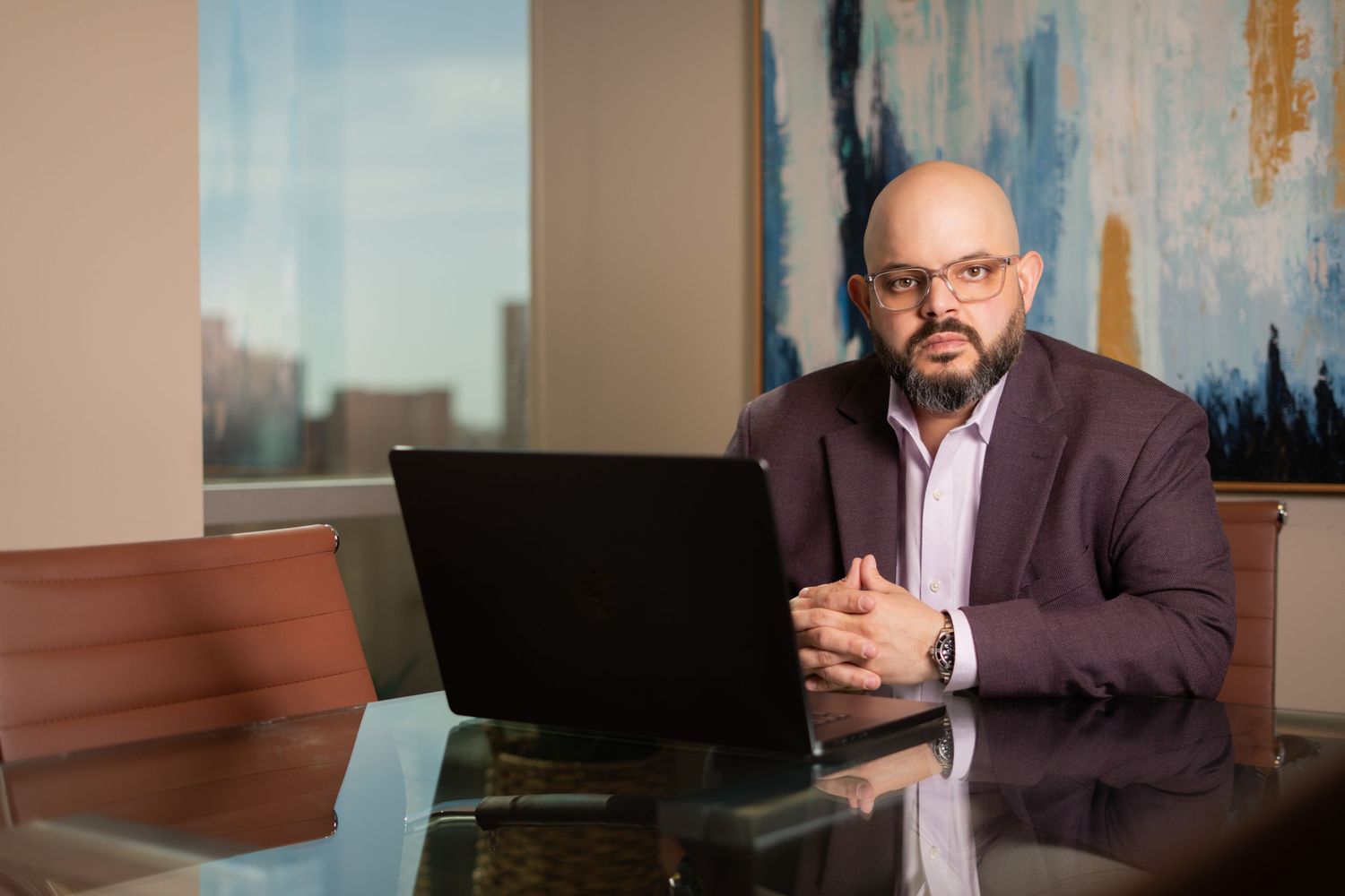 Professional in a brown suit sits at a reflective desk with laptop in modern office setting.