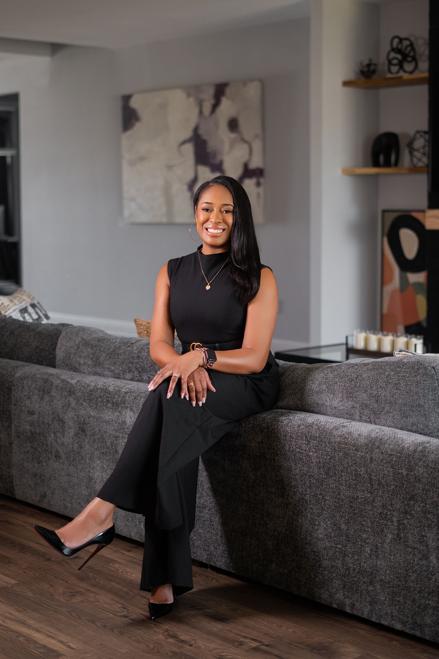 A person in a sleeveless black outfit sits elegantly on a gray couch in a modern living room with abstract wall art.
