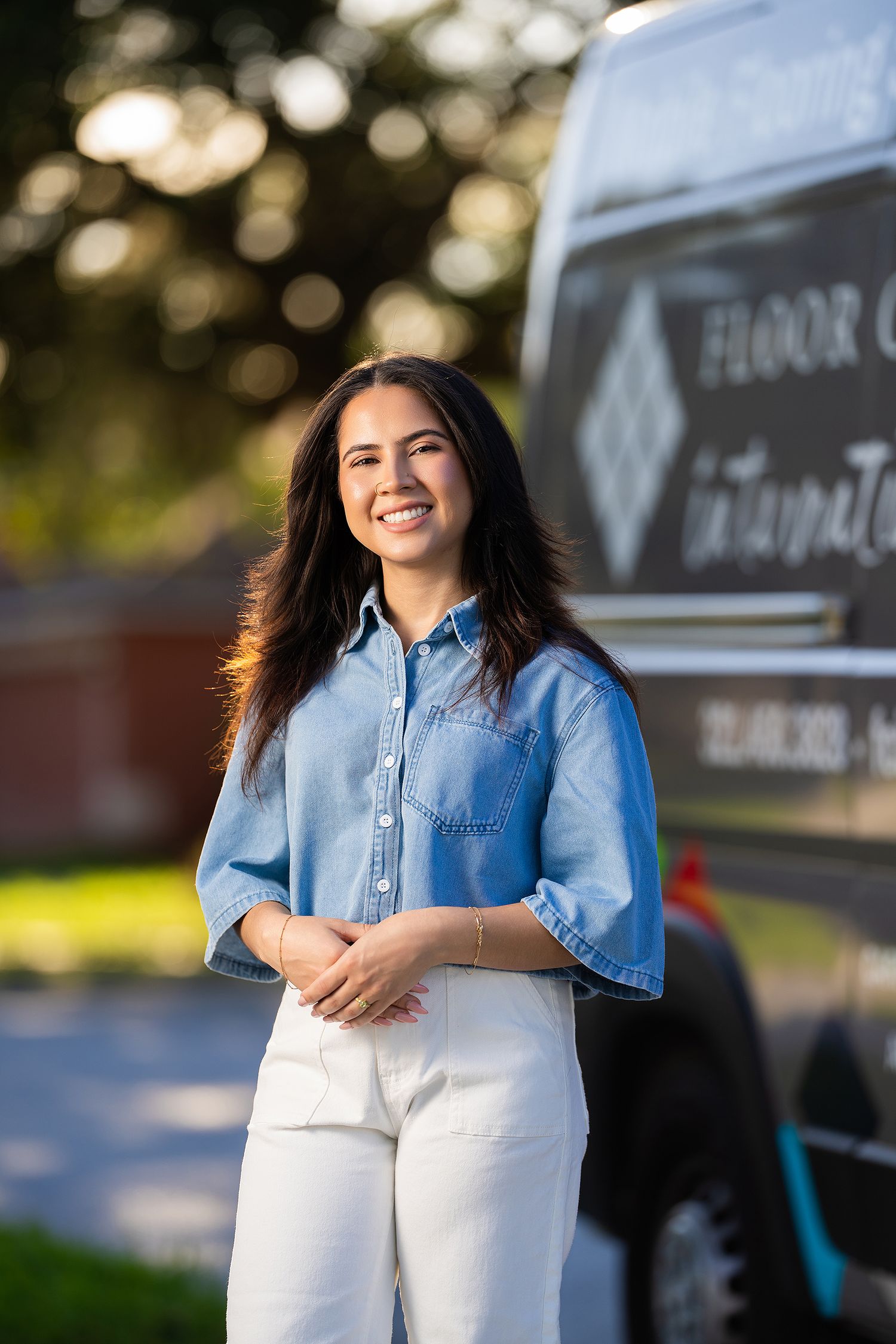 A woman in a chambray shirt and white pants stands confidently against a blurred outdoor background.