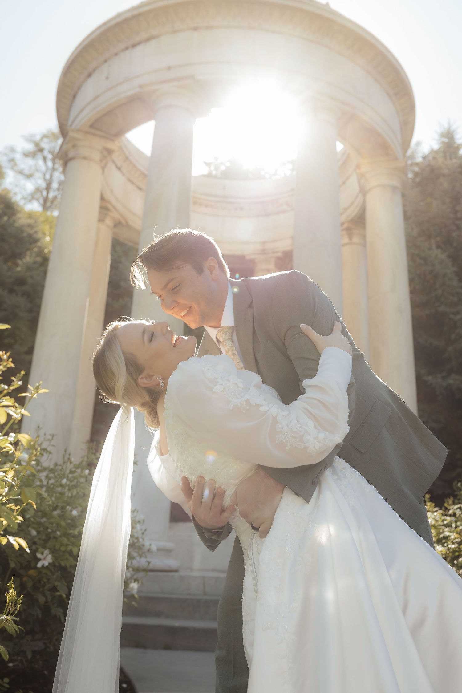 A bride and groom share a romantic dip kiss in front of a sunlit classical stone rotunda on their wedding day.