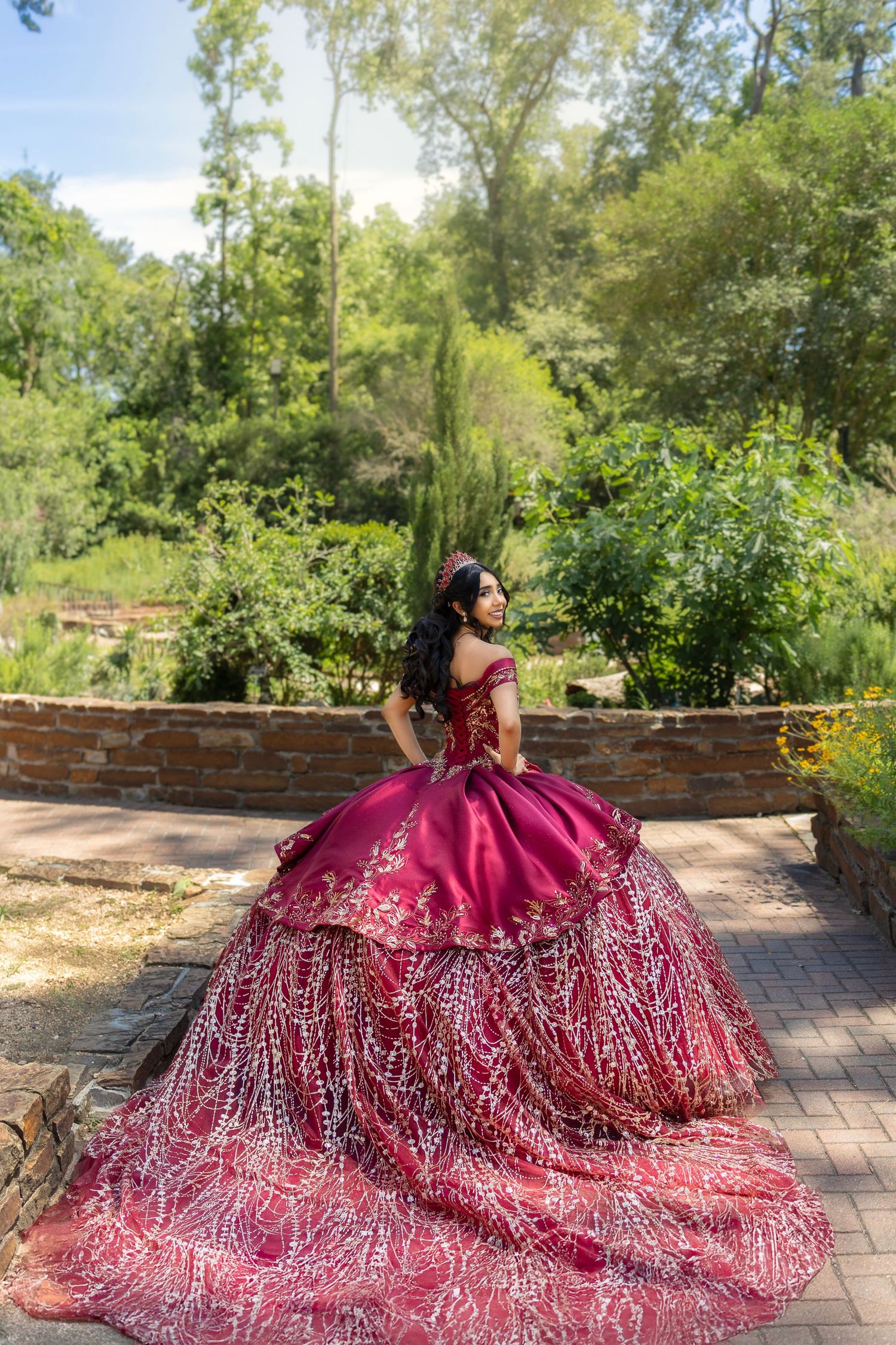 A burgundy quinceañera ball gown with sparkly embellishments flows gracefully across brick pavers in a garden setting.
