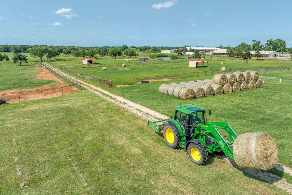 Green John Deere tractor moving hay bales across a rural farm field with red barns visible in the distance.