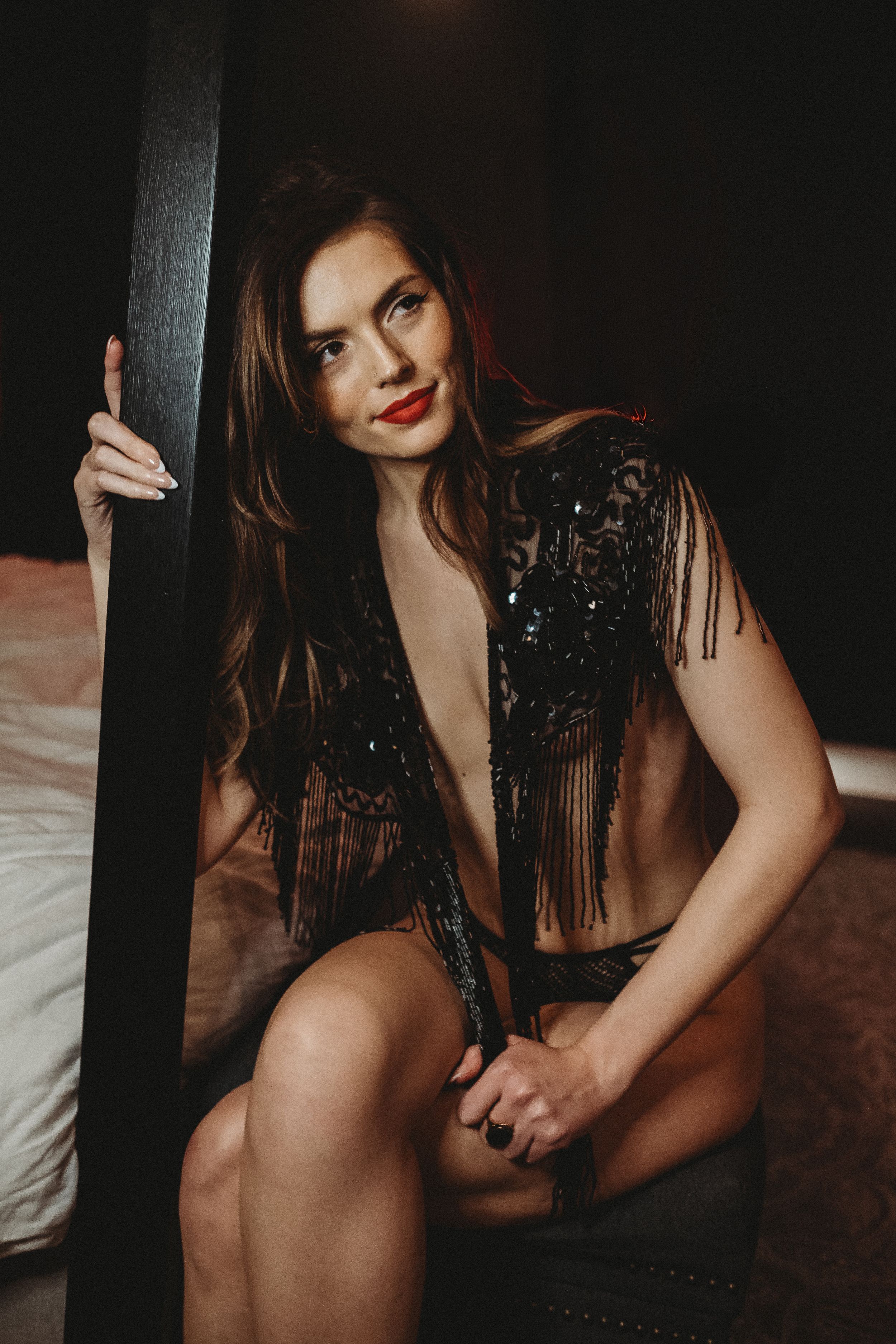 Brunette woman in black sequined fringe top poses beside dark post in moody bedroom setting.