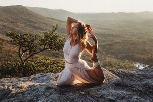 Woman in white outfit practicing yoga pose on a rocky mountaintop at golden hour sunset.