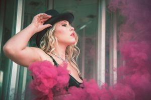 Blonde woman in black lingerie and hat posing dramatically with pink smoke in the background.