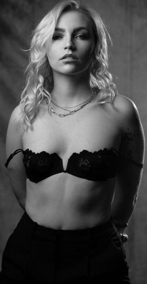 Black and white portrait of a blonde woman in a black lace bralette with layered necklaces and tattoos.
