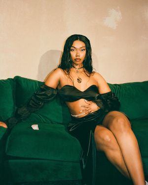 Young woman in a black satin crop top and skirt posing on a green velvet sofa with gold jewelry.
