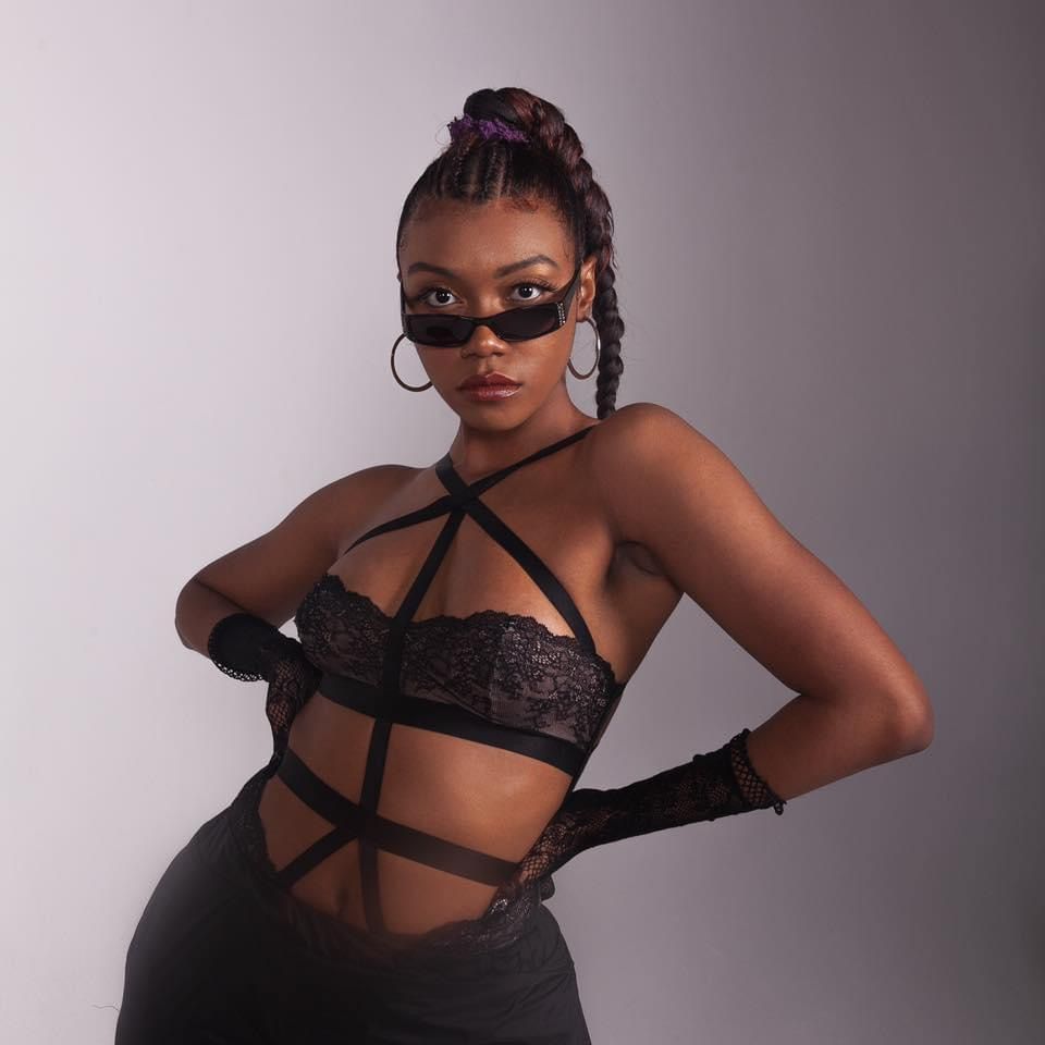 Woman in black strappy lace bralette with sunglasses and gloves poses confidently in studio.
