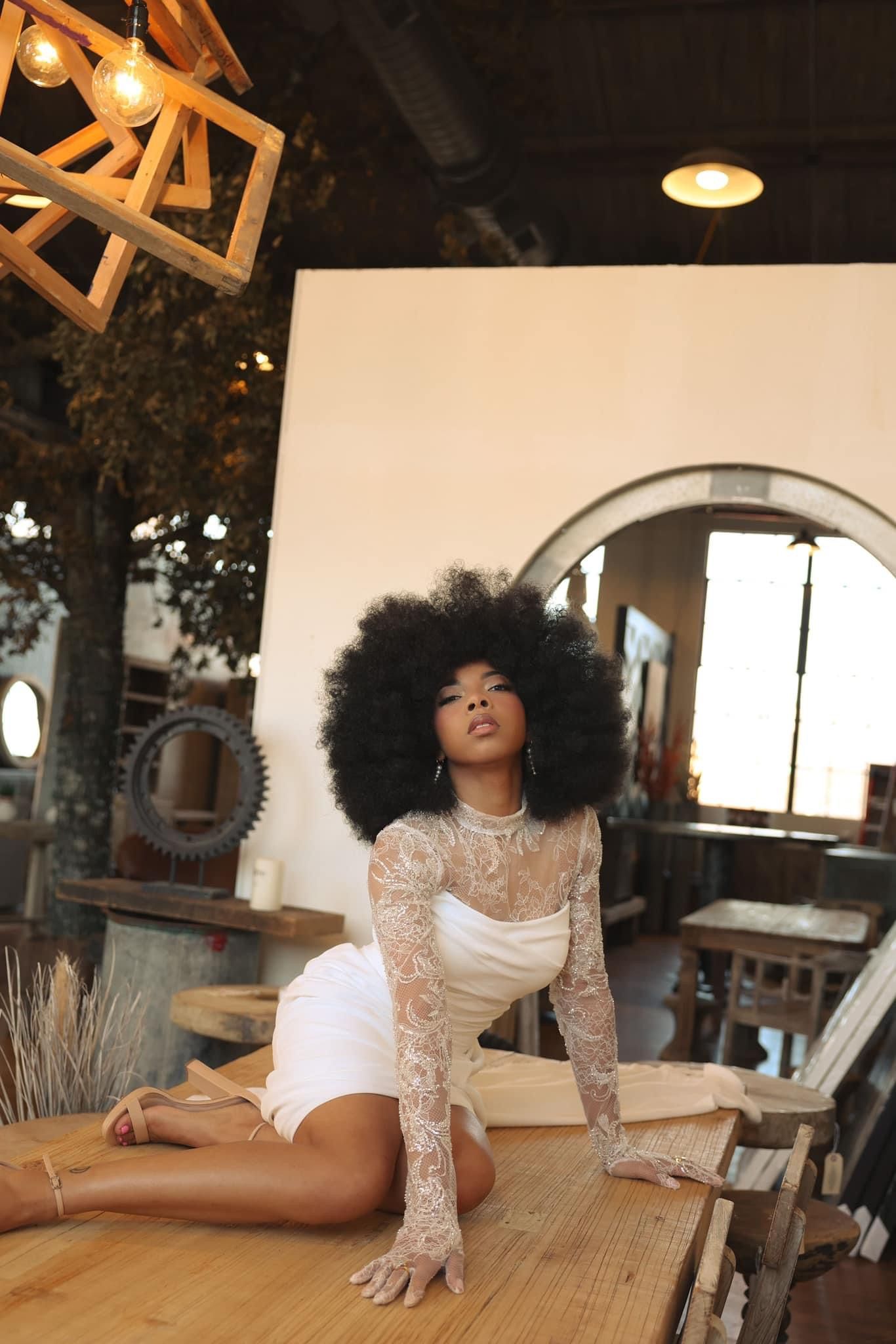 Woman with large afro hairstyle wearing a white lace dress posing on a table in a rustic interior.