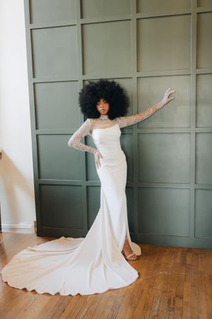 Elegant bride in a white fitted gown with lace sleeves and large afro hairstyle posing by a green wall.