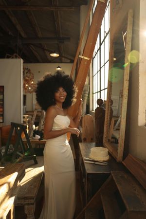 Smiling bride in a white strapless gown with natural afro hair standing in a sunlit rustic warehouse space.