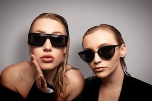 Two stylish women wearing trendy sunglasses posing together against a neutral gray background.