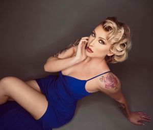 Woman in a blue dress with a rose shoulder tattoo and vintage-style blonde updo posing on the floor.