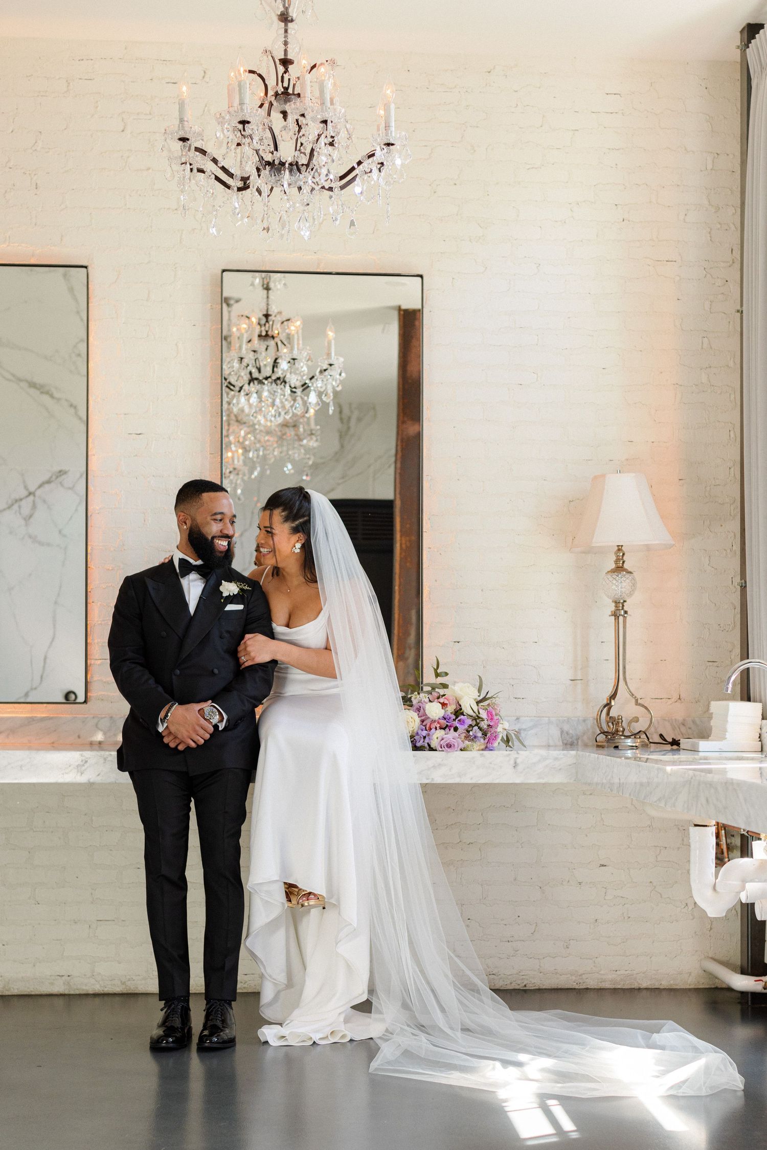 A romantic wedding moment captured under an elegant crystal chandelier with light streaming through a white-painted interior.