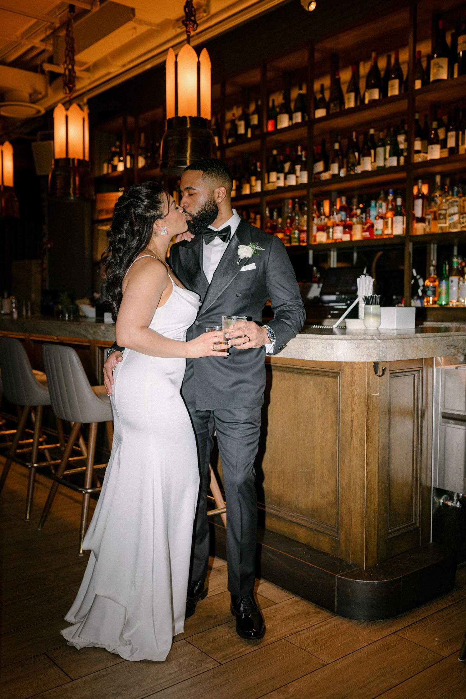 An intimate wedding moment at a classic wooden bar with warm pendant lighting and bottle displays.