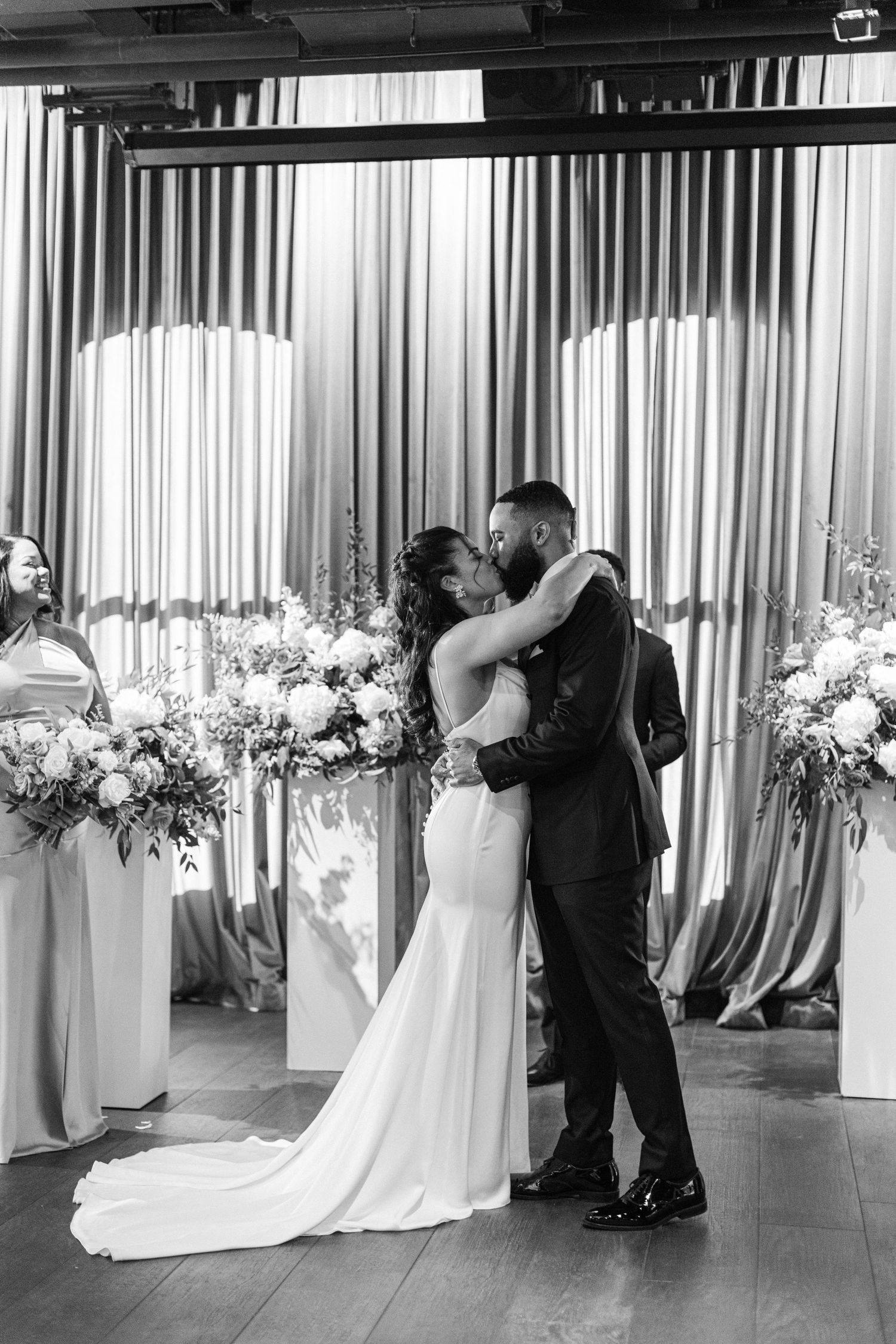 Black and white photo of newlyweds sharing their first kiss at wedding ceremony with floral decor.