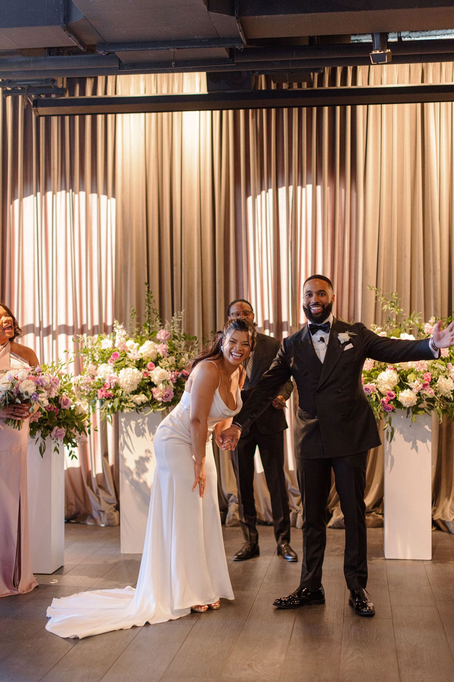 Wedding couple celebrates with guests in a modern venue decorated with floral arrangements and curtains.