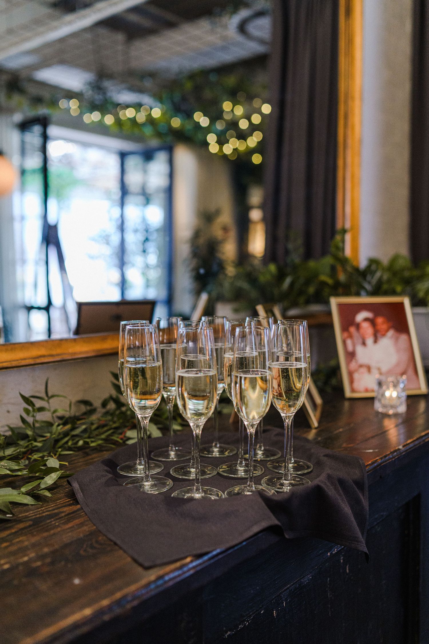 Champagne flutes arranged on wooden table with string lights and greenery in background.