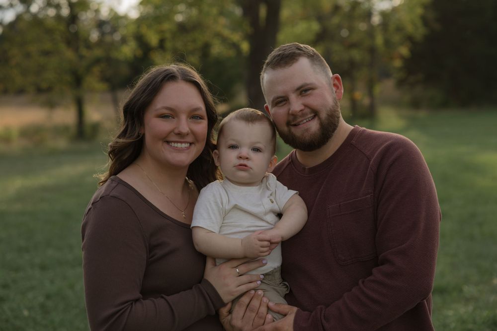 Portrait of a young family wearing matching brown sweaters outdoors on a grassy field.