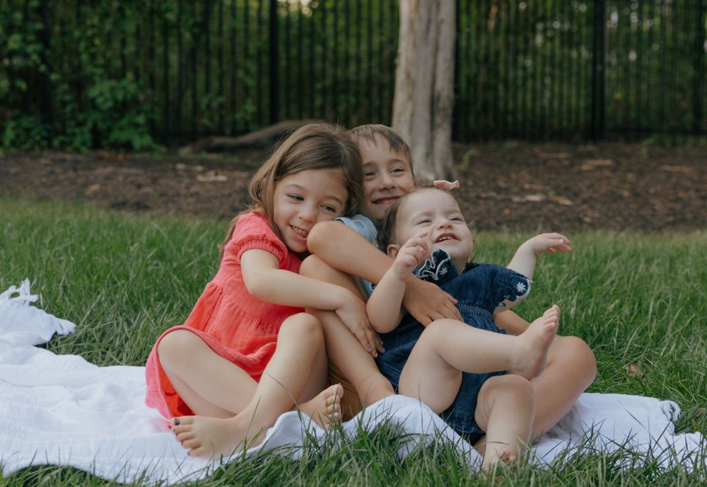 Children hugging and laughing together on white blanket outdoors.