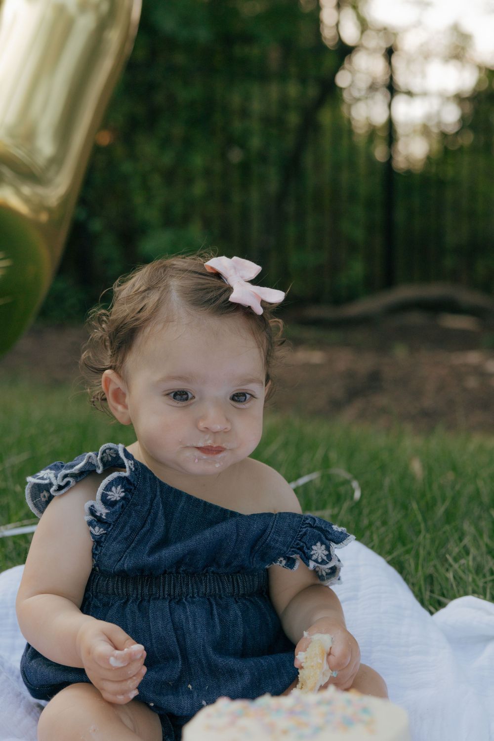Baby in denim dress with bow enjoys first birthday celebration outdoors.