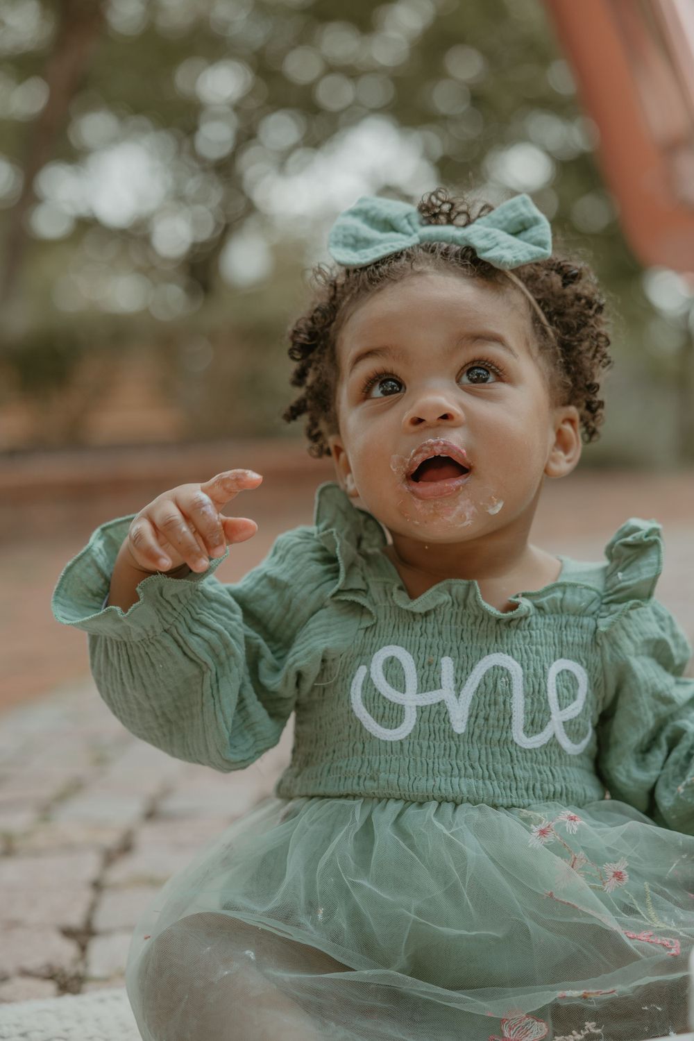 Close up of toddler in mint green dress with bow pointing upward while sitting outside.