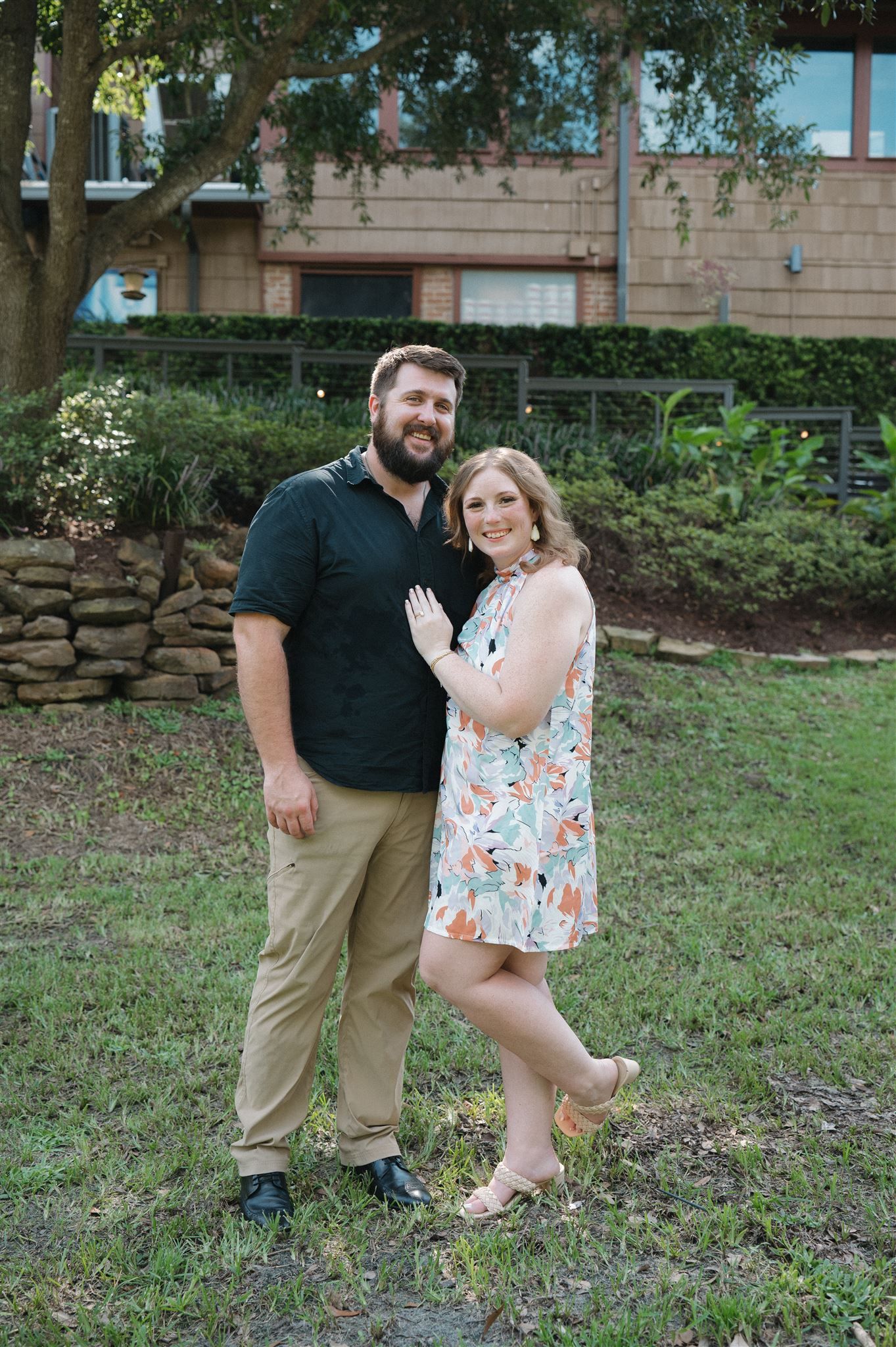 A couple poses outdoors in front of a stone wall and greenery, with one wearing a floral dress and the other in casual attire.